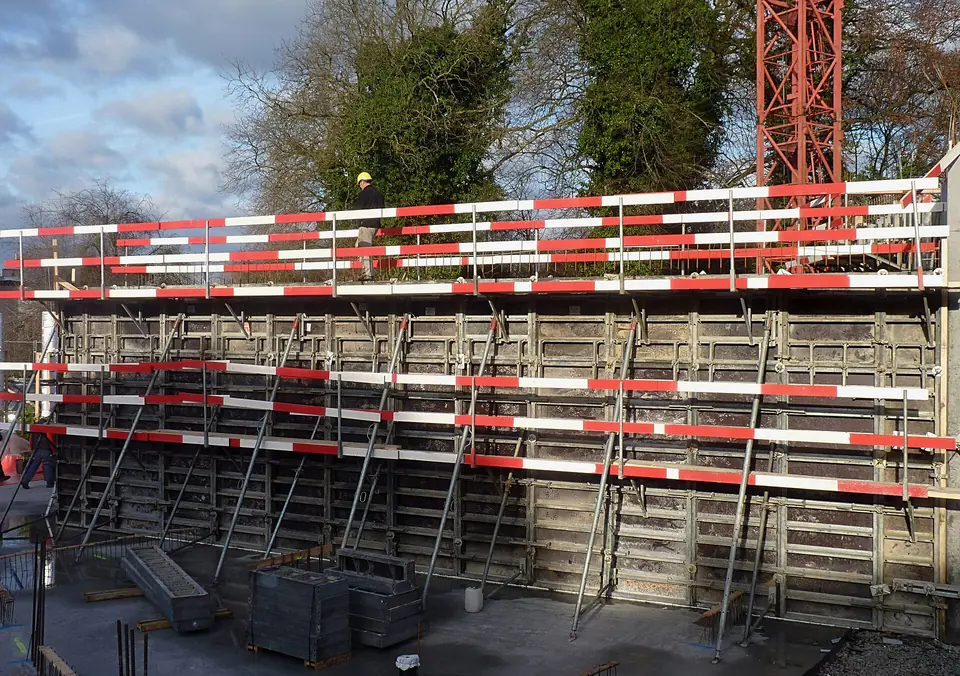 Coffrage de mur en béton sur un chantier en Belgique, avec banches métalliques, passerelles de travail et garde-corps rouges et blancs.