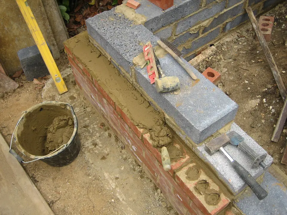 Mur en cours de montage associant briques rouges et blocs de béton, avec truelle, niveau, marteau et seau de mortier posés sur le chantier.